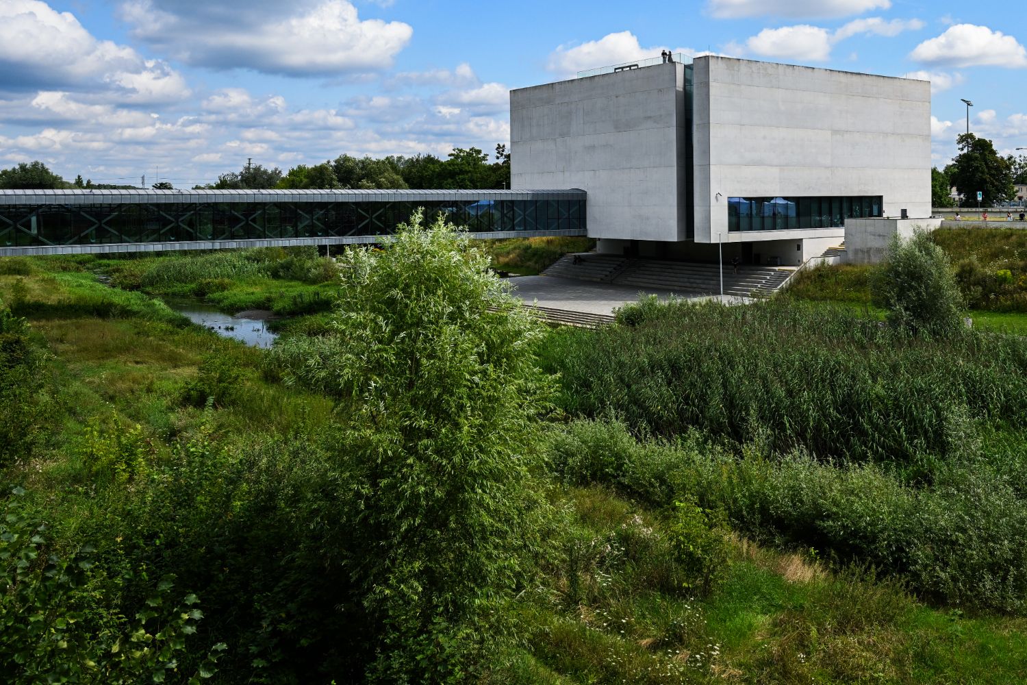 The concrete Gate of Poznań building and its glazed footbridge over the Cybina, surrounded by greenery