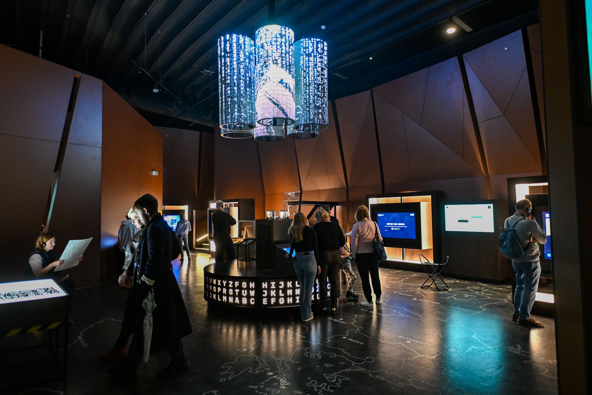 Inside the Enigma Cipher Centre exhibition: a chandelier of glowing letters and visitors at the interactive stations