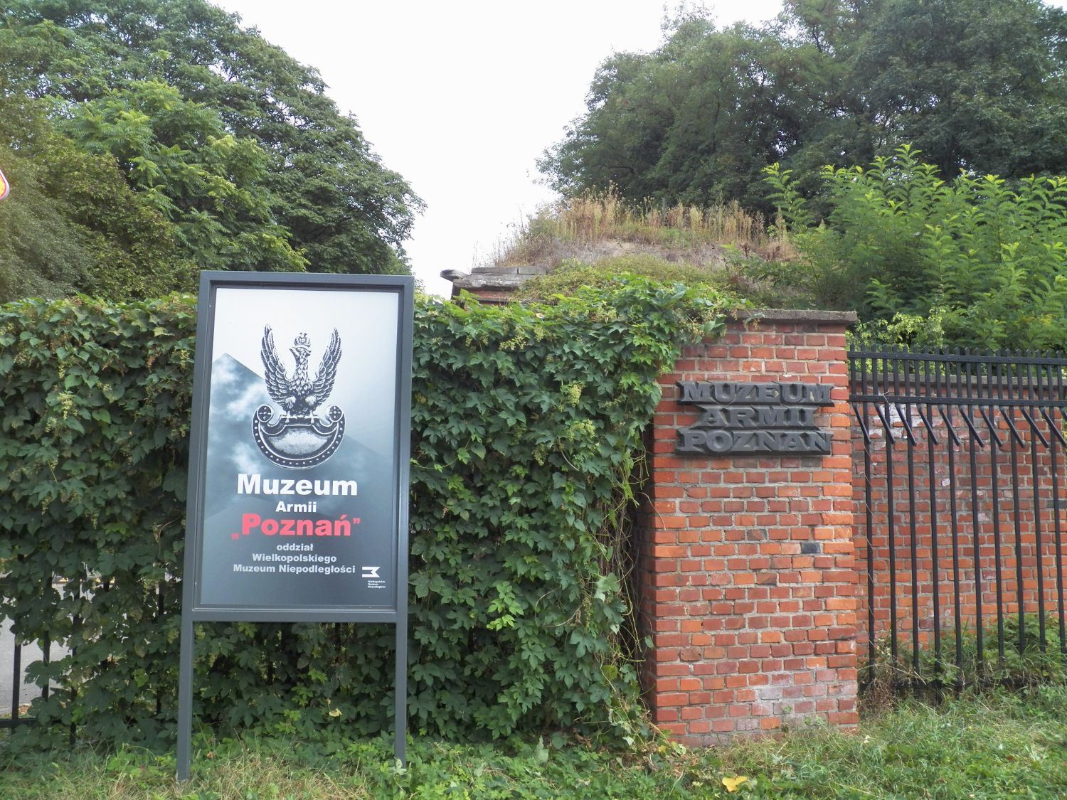 An information board and a brick gatepost with the “Muzeum Armii Poznań” sign, among the greenery of Cytadela Park