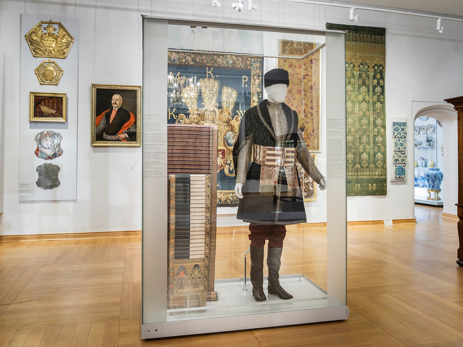 A display case with a mannequin in historic dress, woven sashes and textiles on the walls in the Museum of Decorative Arts